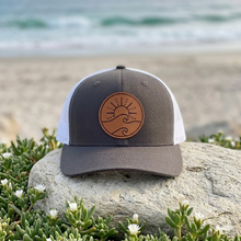 Load image into Gallery viewer, A two-tone trucker hat with a dark gray charcoal front panel and brim, white mesh back, and a brown circular sun-and-wave patch on the front, displayed on a rock with a beach and ocean blurred in the background.