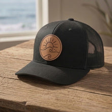 Load image into Gallery viewer, Black cap with a leather patch on a wooden surface with a beach view in the background