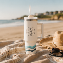 Load image into Gallery viewer, A white handled tumbler with a straw sits on a sandy beach draped with a beige blanket. The tumbler has a black sun-and-wave logo and teal and dark blue wavy stripes near the bottom.