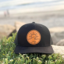 Load image into Gallery viewer, Black cap with tan leather patch logo on grass by a body of water