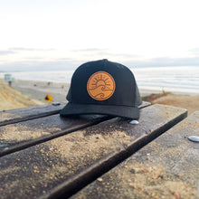 Load image into Gallery viewer, Black cap with a tan leather patch logo on a wooden bench by the beach
