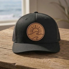 Load image into Gallery viewer, Black cap with a brown leather patch featuring a wave design on a wooden surface with a beach view.
