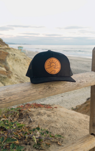 Black trucker hat with a tan leather patch logo on a wooden railing overlooking a beach.