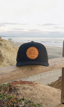 Load image into Gallery viewer, Black cap with a tan leather patch sun waves logo on a wooden railing with a beach background
