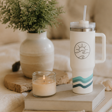 Load image into Gallery viewer, A white handled travel tumbler with a lid and straw stands on stacked books beside a lit candle and a vase with greenery, set on a soft beige sofa.
