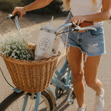 Load image into Gallery viewer, A woman riding a light blue bicycle on a sunlit road, carrying a white insulated mug in a wicker basket filled with white flowers. She wears a white sleeveless top and denim shorts.
